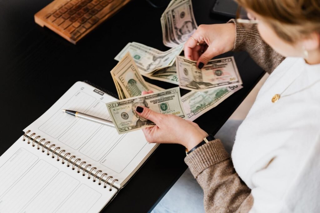 Woman organizing finances, counting dollar bills at desk with open notebook.
