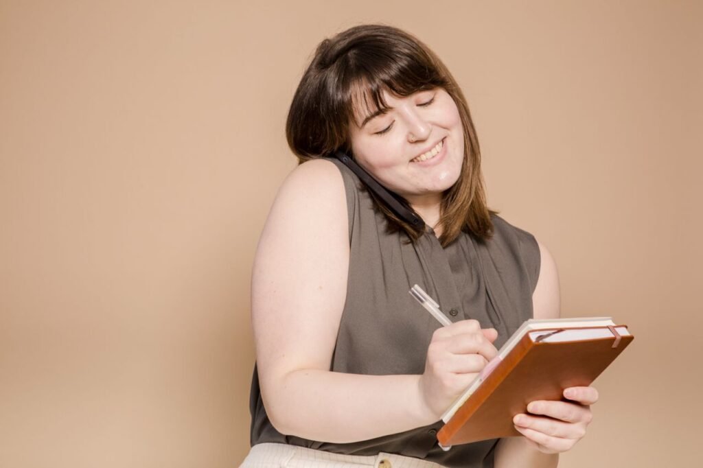 Positive Asian female with pen smiling and taking notes in notepad while having phone conversation on beige background in light studio